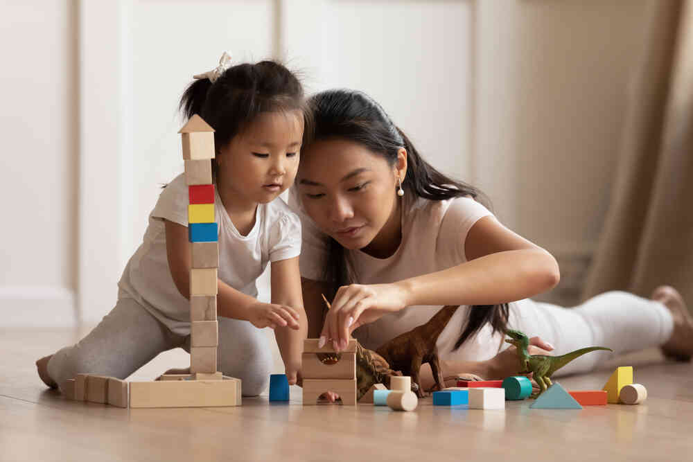 A woman and a young girl lay on the floor, playing with wooden blocks. The girl watches intently as the woman stacks the blocks, with a colorful tower nearby. The scene is bright and filled with a sense of focused playtime and nurturing child care.